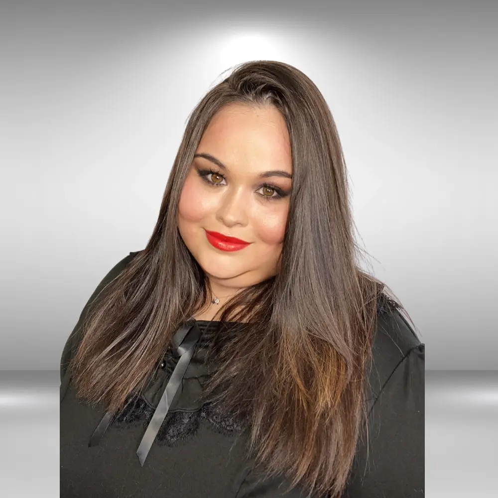 A person with long, straight brown hair and bold makeup, including red lipstick and dark eye makeup, smiles slightly at the camera. They are wearing a black top with a ribbon detail against a light gray background, exuding an influencer’s charm with runway-ready talent.